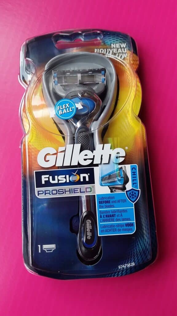 A Gillette Fusion ProShield Chill razor in its packaging, displayed on a bright pink surface. The packaging shows the razor with a blue and silver handle and highlights features like “FlexBall” and lubrication before and after the blades.