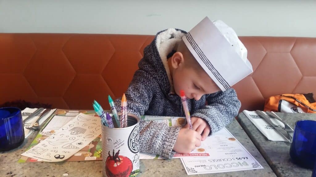 A young child wearing a paper hat and grey jumper leans over a restaurant table, colouring a menu with crayons and felt-tip pens. The table has activity sheets, a cup of pens, blue glasses, and neatly arranged cutlery in serviettes.