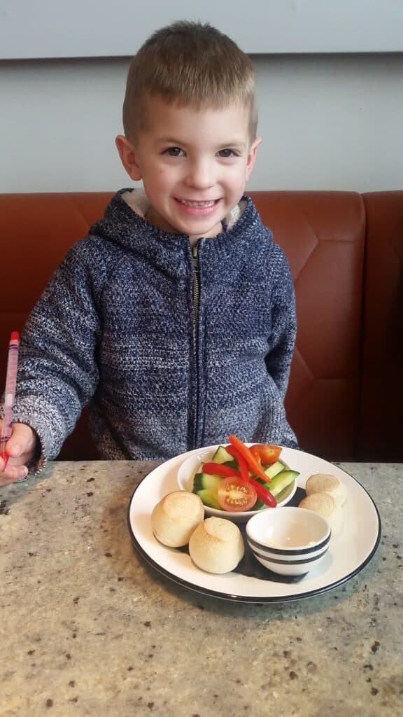 A young boy with short hair and a blue zip-up jumper smiles while sitting at a table. In front of him is a plate with round bread rolls, a small bowl of dip, and sliced vegetables including cucumber, tomato, and peppers.