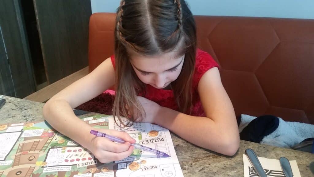 A young girl with brown hair in plaits sits at a granite table, focused on solving a colourful puzzle or activity sheet with a purple pen. She wears a red top and rests one arm on the table. Cutlery is placed to her right on a serviette.