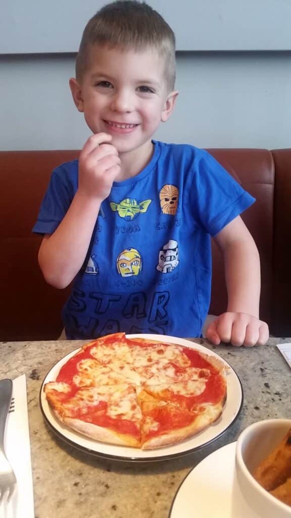 A smiling young boy with short brown hair sits at a table in a restaurant, wearing a blue Star Wars T-shirt. He is about to eat a cheese pizza on a white plate in front of him. A fork, knife, and a bowl are also on the table.