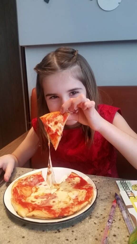 A young girl with light brown hair, wearing a red dress, sits at a table. She smiles and lifts a cheesy slice of pizza from a plate, stretching melted cheese. The table has colourful pencils and a sheet of paper nearby. The background features a brown bench and light-coloured wall.