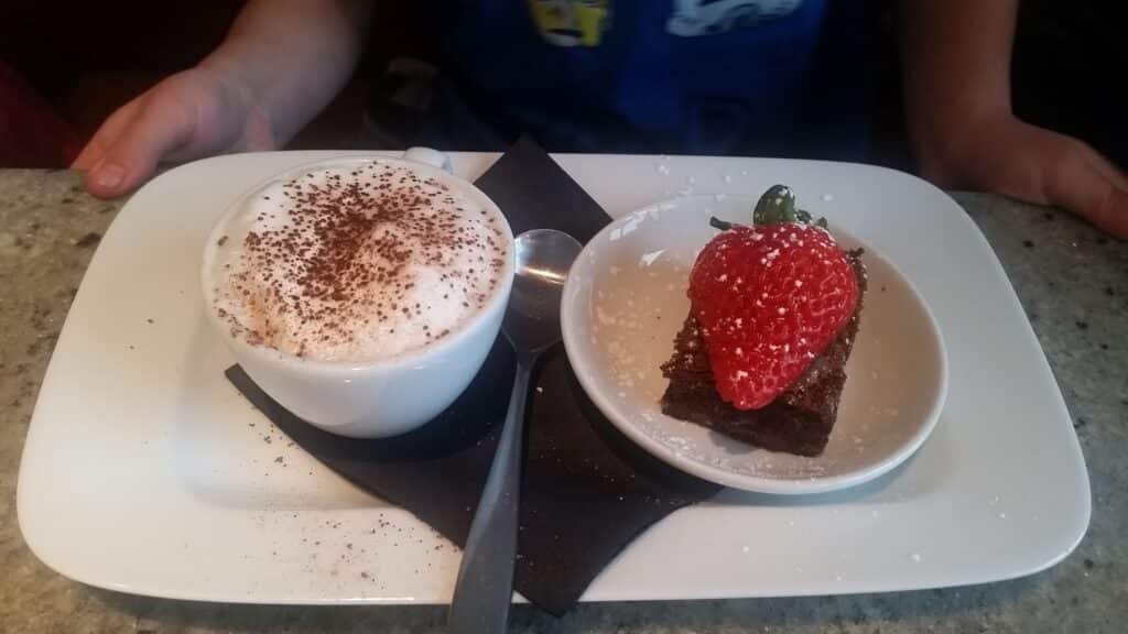 A white saucer holds a foamy cappuccino dusted with cocoa, a spoon, and a chocolate brownie topped with a large strawberry and icing sugar, all on a black serviette, with a child's hands visible in the background.