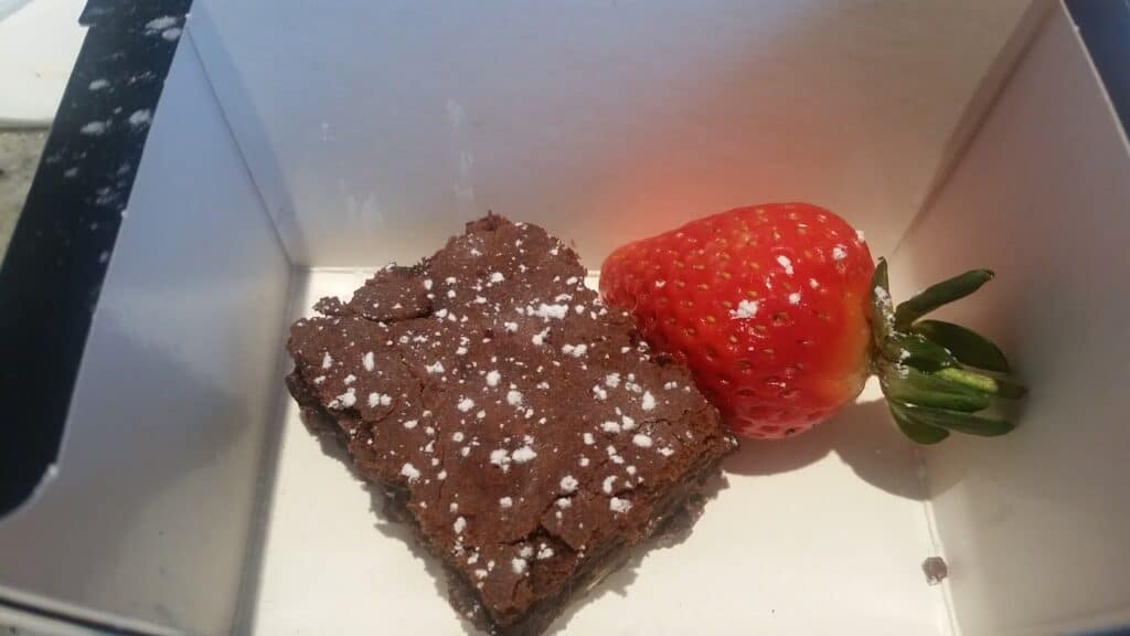 A single chocolate brownie dusted with icing sugar sits in a white box next to a whole red strawberry with a green leafy top. The box has dark edges and there is some sunlight shining onto the treats.