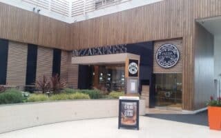 The image shows the exterior of a PizzaExpress restaurant. The building features modern wood panelling, large windows, and the PizzaExpress logo above the entrance. There are green plants in front, and a sign advertises pizza offers near the doorway.