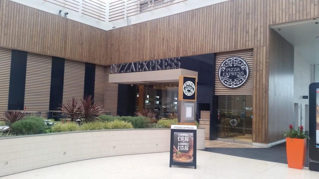 The exterior of a PizzaExpress restaurant features wooden panelling, large windows, modern landscaping with plants, and two signs with the PizzaExpress logo. A freestanding menu board displays special offers near the entrance.