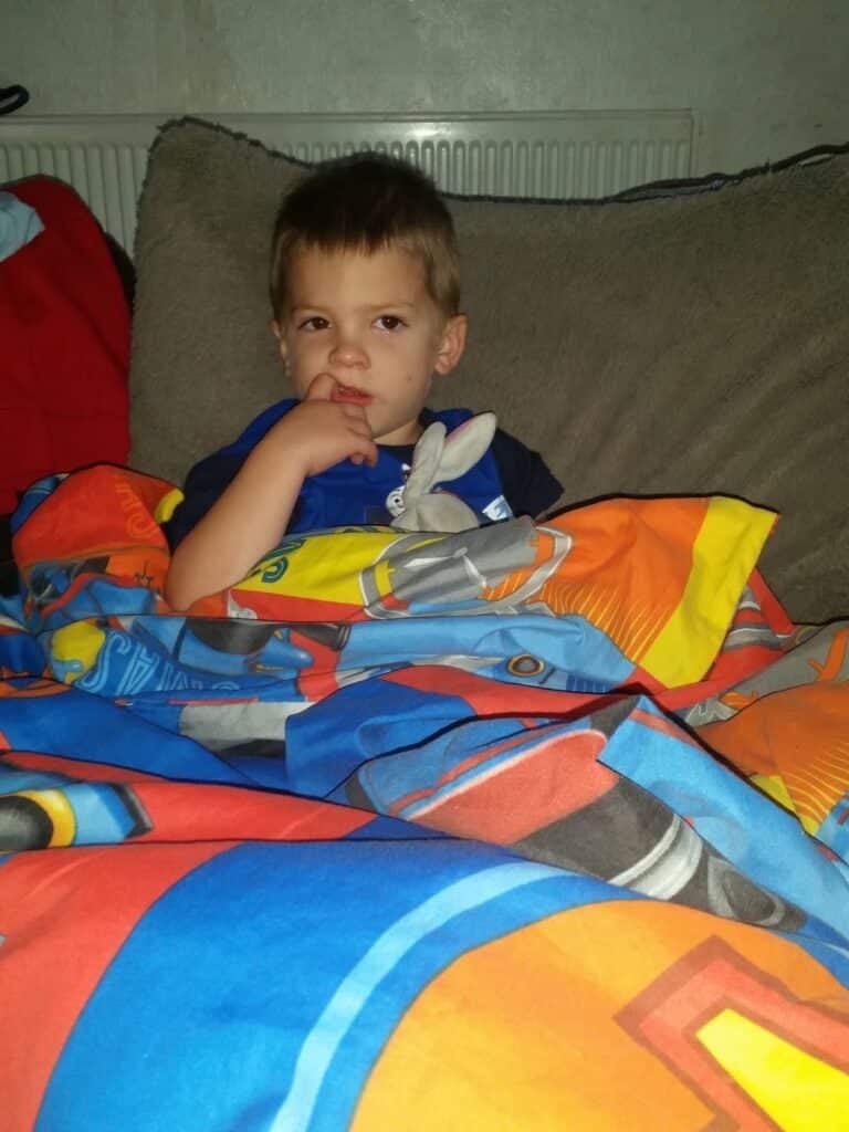 A young boy sits on a bed covered with a colourful cartoon-themed duvet. He is sucking his thumb, holding a small grey soft toy, and looking off to the side with a thoughtful expression. The background shows a pillow and a radiator.