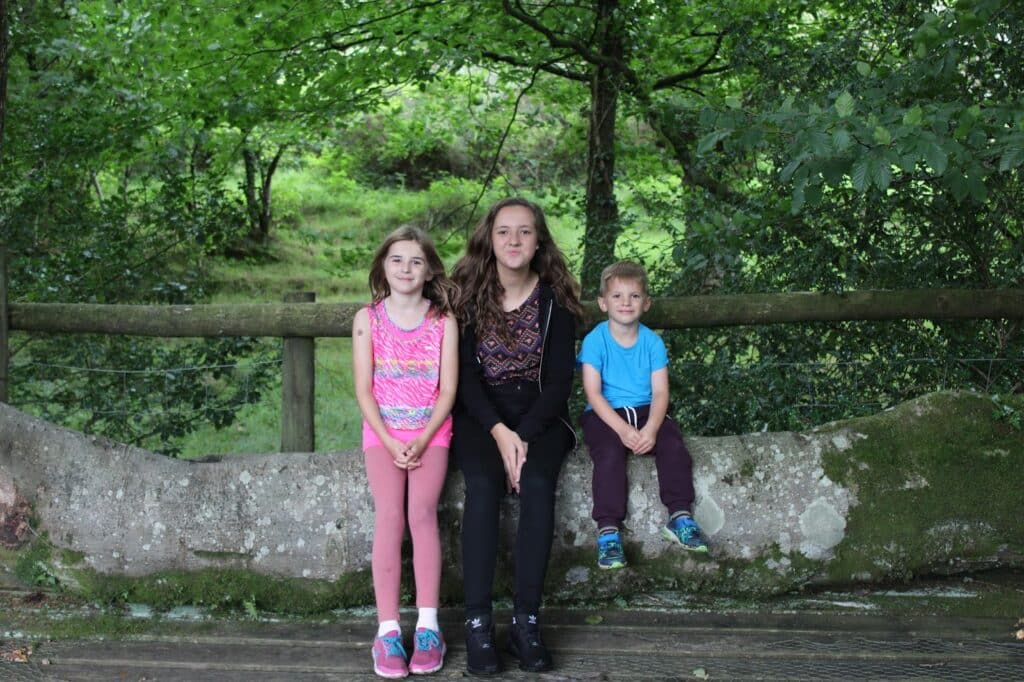 Three children sit on a large, mossy tree trunk in front of a wooden fence. Behind them is lush green foliage. The girl on the left wears pink clothes, the girl in the centre wears black, and the boy on the right wears a blue shirt and maroon trousers.