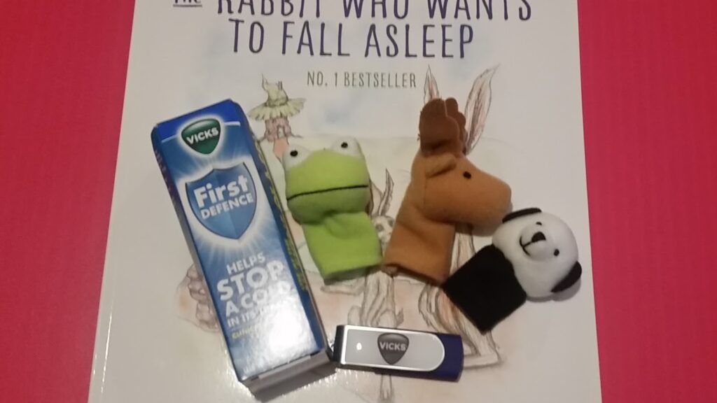 A book titled “The Rabbit Who Wants to Fall Asleep” lies on a pink surface. On top are a Vicks First Defence nasal spray box, three finger puppets (a green frog, brown horse, black-and-white panda), and a USB stick labelled “Vicks”.