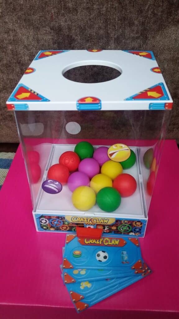 A toy claw machine with a clear box contains colourful plastic balls—red, yellow, green, and purple—on a white base labelled "Crazy Claw." Instruction sheets with game graphics lie on a pink surface in front of the machine.