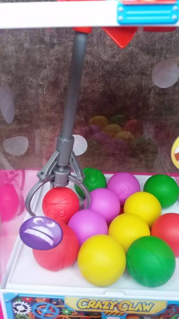 A claw machine's metal claw grips a red ball with a frowning face sticker. Inside the machine are colourful plastic balls—yellow, green, pink, and red—on a white base. The label "CRAZY CLAW" is visible at the front bottom of the machine.