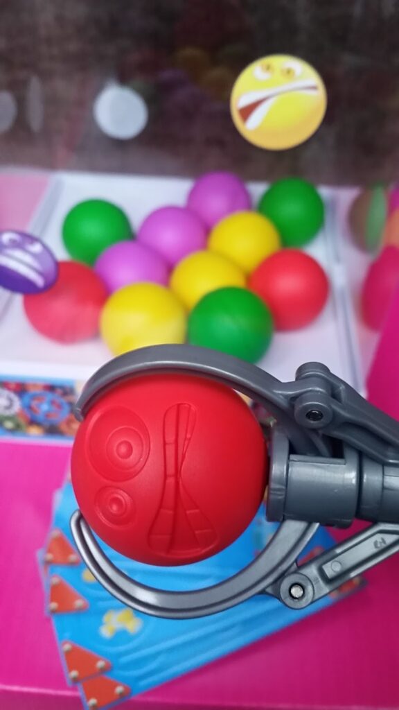A claw machine grips a red plastic ball with a surprised face design. Behind it are yellow, pink, green, and purple balls. The background is pink, featuring playful emoji and science flask graphics, with multicoloured game cards visible at the bottom.