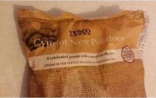 A bag of Tesco Cypriot New Potatoes with brown, rustic packaging. The label reads: “A celebrated potato with a sweet earthy taste. Grown in the fertile red soils of southern Cyprus.” Part of the netted bag and potatoes are visible.
