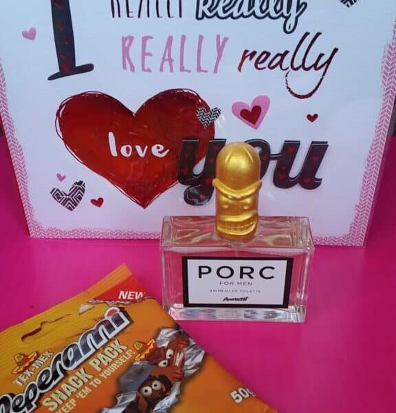 A gift set on a pink surface features a card reading “I really really really love you” with hearts, a bottle of PORC perfume with a gold pig-shaped cap, and a pack of Peperami Snack Pack with cartoon sausages on the packaging.