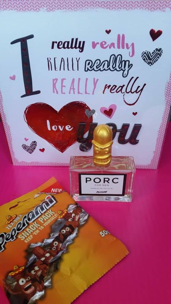 A card with the phrase "I really really really love you" in pink and red fonts stands behind a perfume bottle labelled "PORC" with a gold cap shaped like a pig's head. In front is an orange pack of Peperami Snack Pack, featuring cartoon sausage characters.