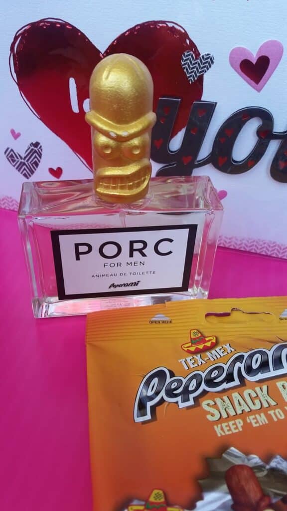 A perfume bottle labelled "PORC FOR MEN" with a gold, cartoonish, angry-faced cap sits on a pink surface. Behind it is a card with hearts and "You" in glitter. In front, a partially visible bag of Texas Pepperell snack sticks appears.