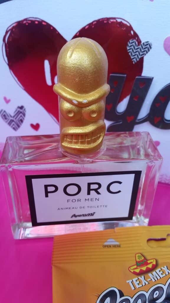 A clear glass bottle labelled "PORC FOR MEN" with a gold, cartoonish figure as the cap. In the background, there's a large red heart and decorative text. At the bottom right, part of a yellow snack packet is visible.