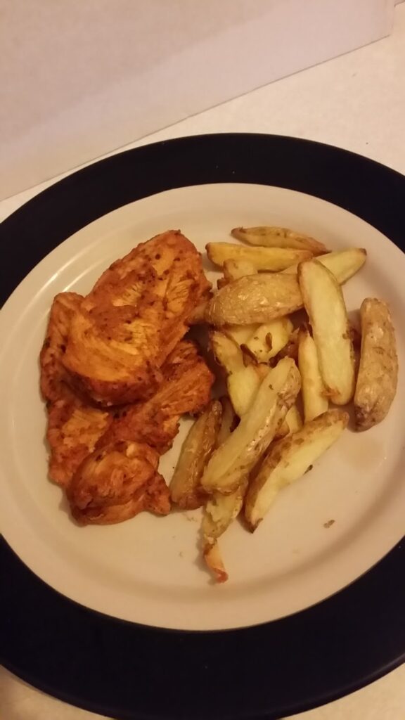 A plate with several pieces of seasoned, cooked chicken breast and a serving of golden, oven-baked potato wedges, arranged side by side on a round white plate with a dark rim. The background is neutral and uncluttered.