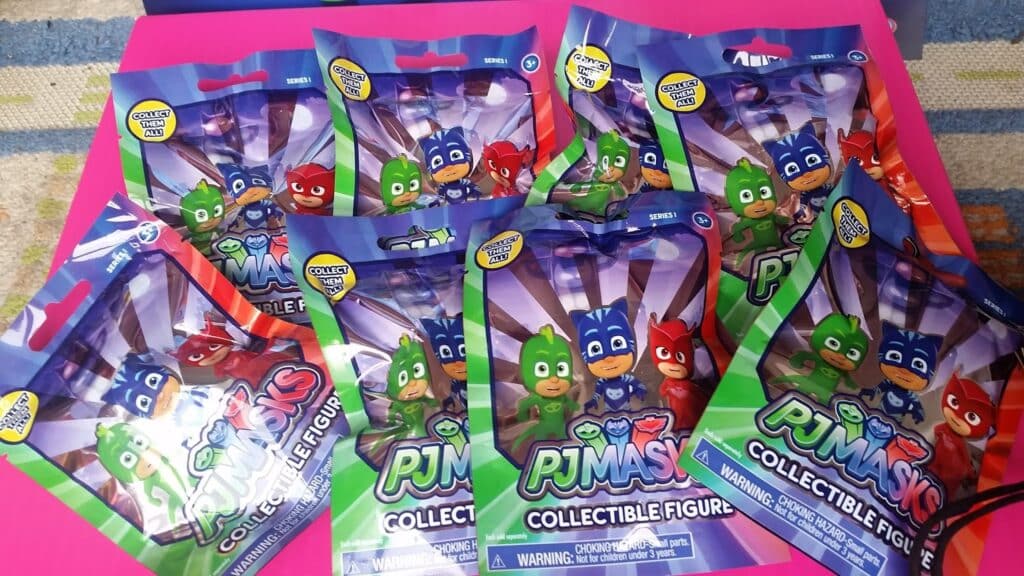 Eight unopened PJ Masks Collectable Figure blind bags are arranged on a bright pink surface. The packaging shows the three main PJ Masks characters—Gekko, Owlette, and Catboy—smiling, with "PJ Masks Collectable Figure" written on each bag.