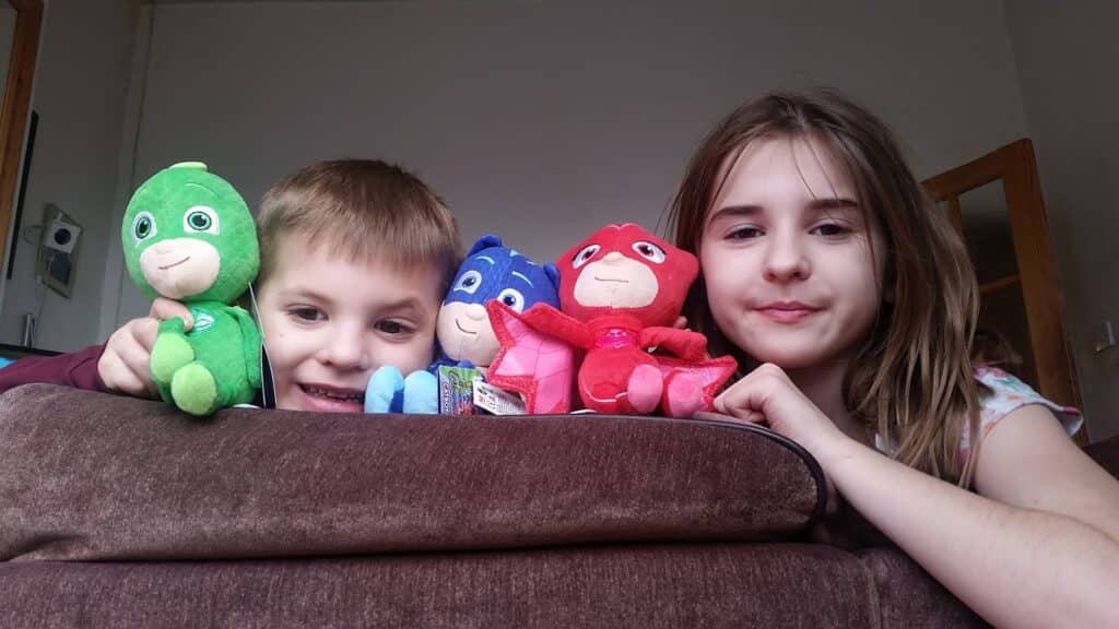 A young boy and girl smile over the back of a brown sofa, each holding colourful PJ Masks plush toys: Gekko (green), Catboy (blue), and Owlette (red). The room is softly lit, with beige walls and a wooden door in the background.