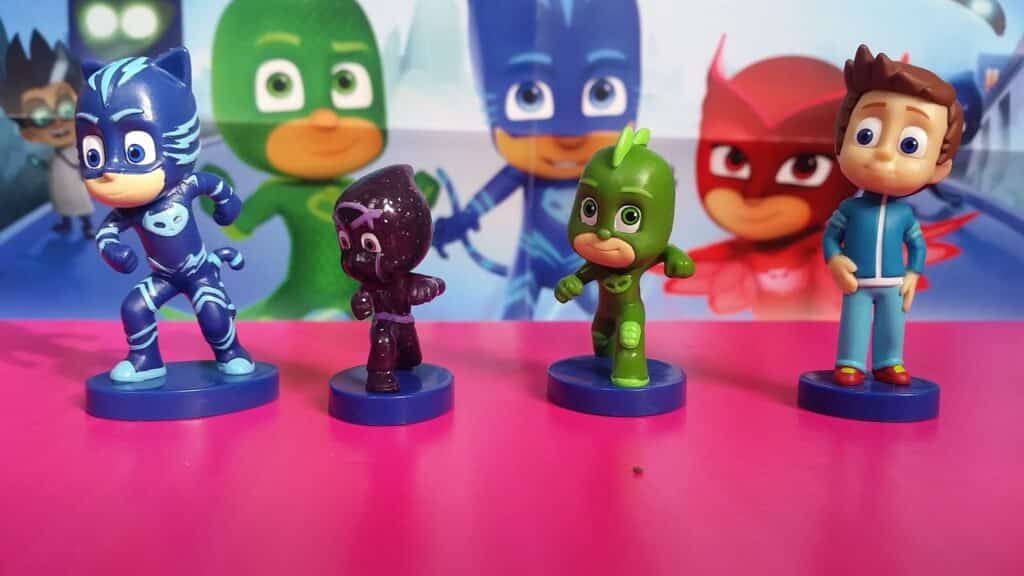 Four PJ Masks character figurines—Catboy, Night Ninja, Gekko, and a boy in blue pyjamas—stand on blue bases on a pink surface, with a colourful PJ Masks-themed background featuring larger character illustrations.