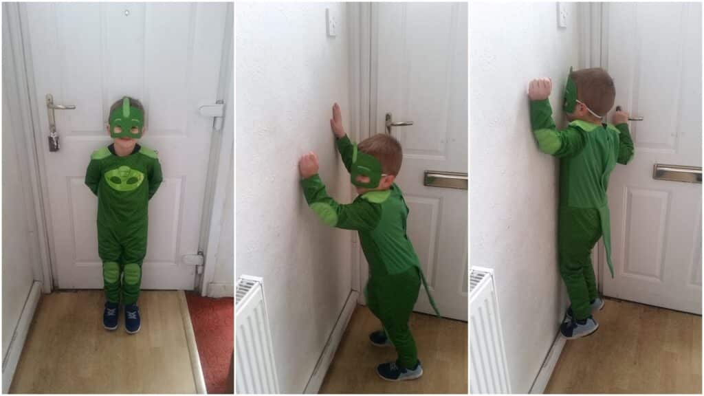 A young child wearing a green superhero costume, mask, and blue shoes stands by a white door. In the first image, they face forward; in the next two, they face and lean toward a corner, mimicking being in time-out. The scene is indoors with white walls and a radiator.