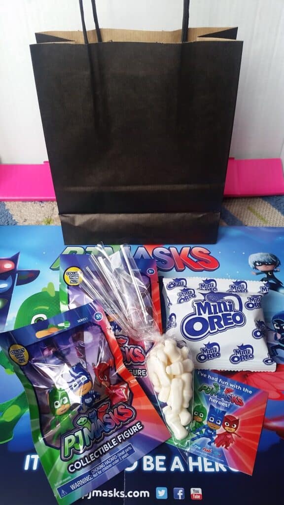 A black paper gift bag sits behind a colourful PJ Masks-themed mat. In front are two PJ Masks collectable figure packets, a clear sweet bag with a twist tie, a pack of Mini Oreos, and some white sweet pieces. Bright, playful colours dominate the scene.