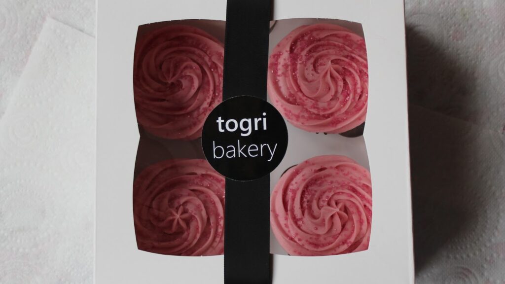 A white bakery box with a black ribbon and a round sticker reading "togri bakery" displays four cupcakes topped with pink, swirl-patterned icing, visible through a clear window. The box rests on a white, textured kitchen roll.