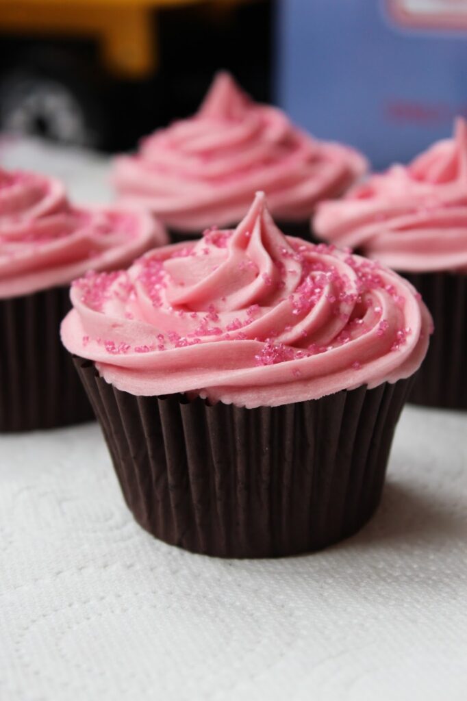 A chocolate cupcake with a dark brown wrapper is topped with a swirl of pink icing and sprinkled with pink sugar crystals. Three more similarly decorated cupcakes are partially visible in the background on a white textured surface.