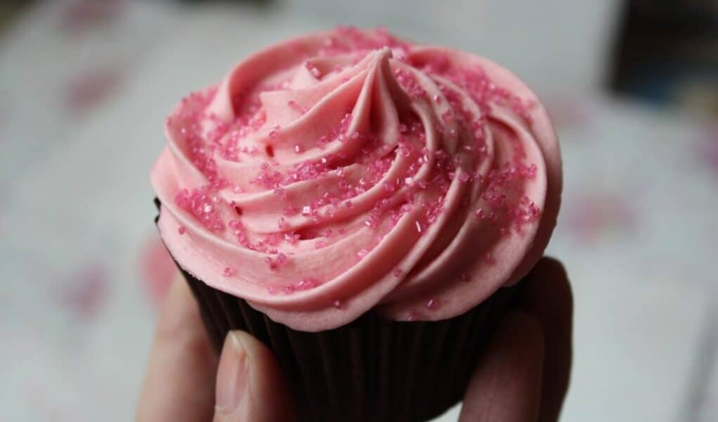 A hand holds a chocolate cupcake topped with a swirl of pink icing and sprinkled with pink sugar crystals. The background is blurred, drawing focus to the cupcake’s decorative texture and vibrant colours.