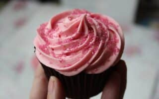 A hand holds a chocolate cupcake topped with a swirl of pink icing and sprinkled with pink sugar crystals. The background is blurred, drawing focus to the cupcake’s decorative texture and vibrant colours.