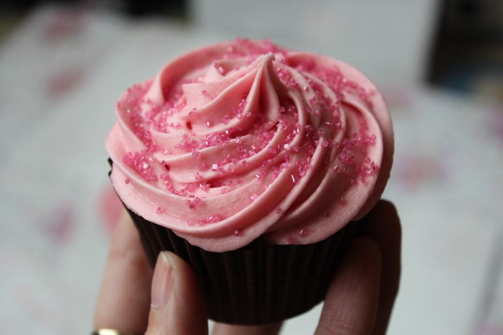 A hand holds a chocolate cupcake topped with pink swirled icing and sprinkled with pink sugar crystals. The background is blurred, drawing focus to the cupcake’s detailed, creamy texture and vibrant colour.