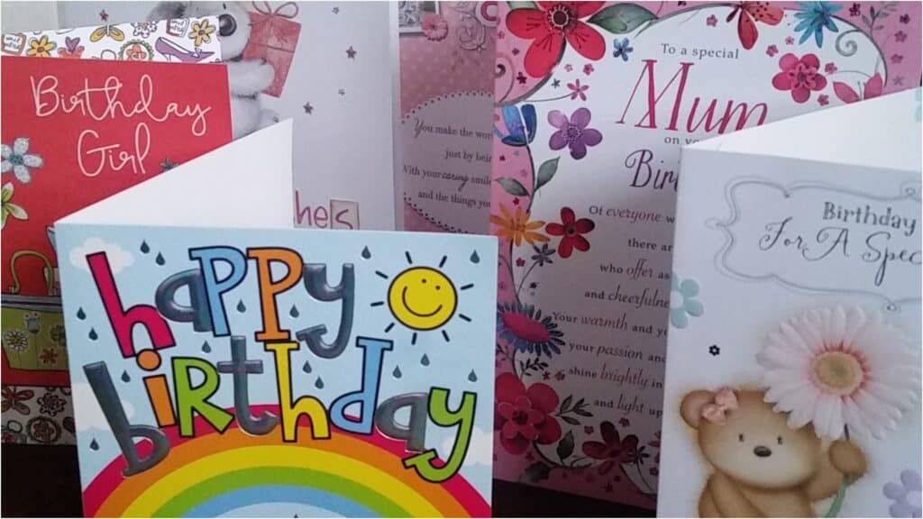 A group of birthday cards stands upright. The cards feature bright designs: a rainbow, a teddy bear with a flower, floral patterns, and the words “happy birthday,” “Birthday Girl,” “Mum,” and “Birthday for a special.”.