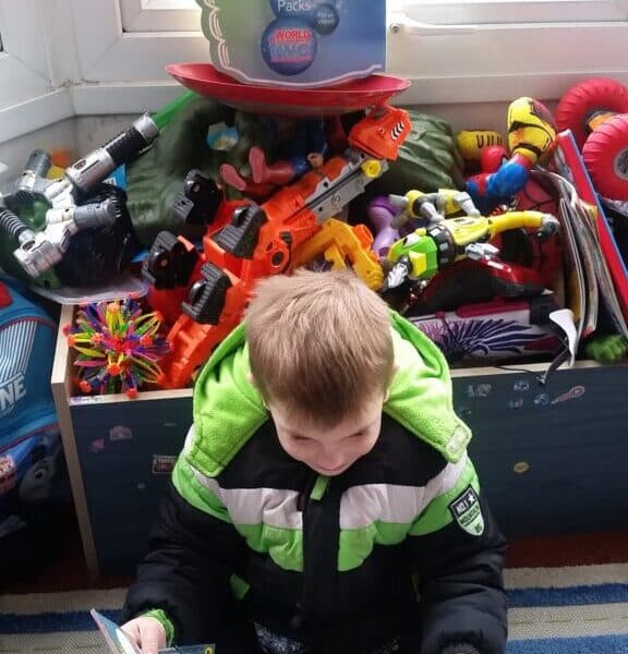 A young boy wearing a black and green winter coat sits on a colourful rug reading a magazine. He is in front of a large toy box filled with various brightly coloured toys and action figures by a window with a “Rocket Packs” display sign on top.