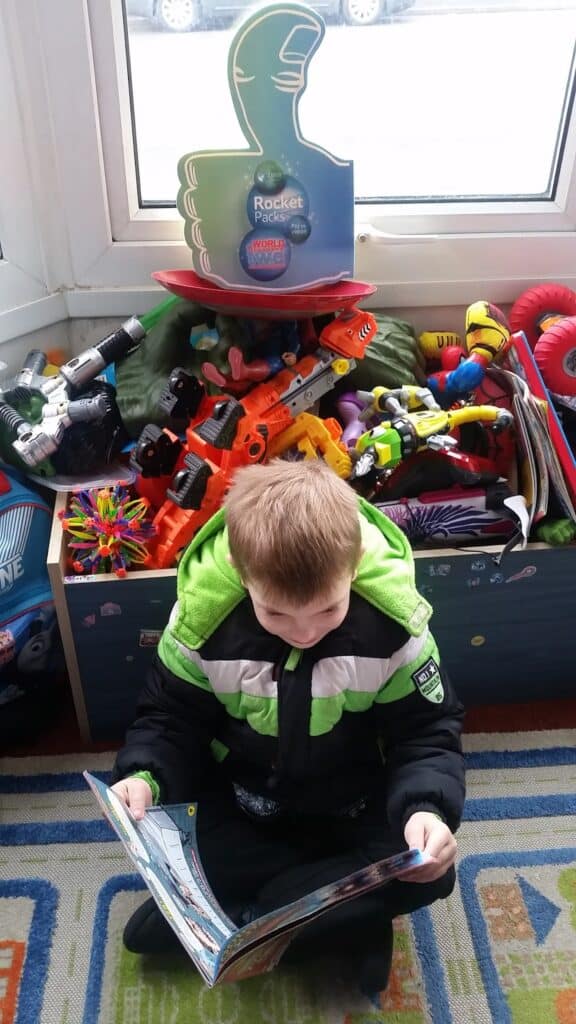 A young boy in a green and black jacket sits cross-legged on a colourful rug, reading a magazine. Behind him is a box overflowing with various toys, including action figures, robots, and balls, near a window with a “Rocket Packs” sign above.