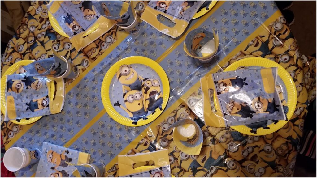 A round table covered with a Minions-themed tablecloth, plates, cups, serviettes, and party favour bags. The plates and party items feature images of yellow Minions characters from the “Despicable Me” films, arranged for a children’s party.