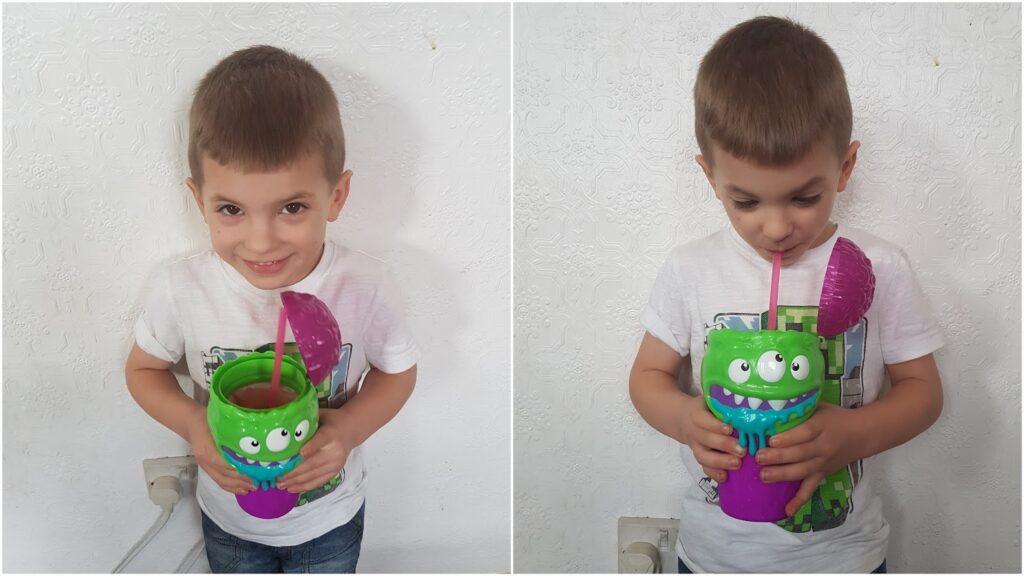 A young boy with short brown hair, wearing a white T-shirt and jeans, stands against a white textured wall. He is holding and drinking from a large, colourful cup with a green monster face, a purple lid, and a pink straw, smiling in the first photo.