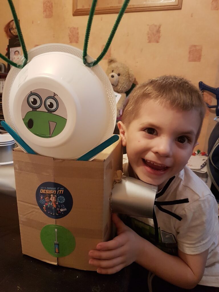 A smiling young boy hugs a homemade robot made from a cardboard box, plastic bowls, and green pipe cleaners. The robot has a cartoon face with big eyes and a sticker on its chest. A teddy bear and other items are in the background.