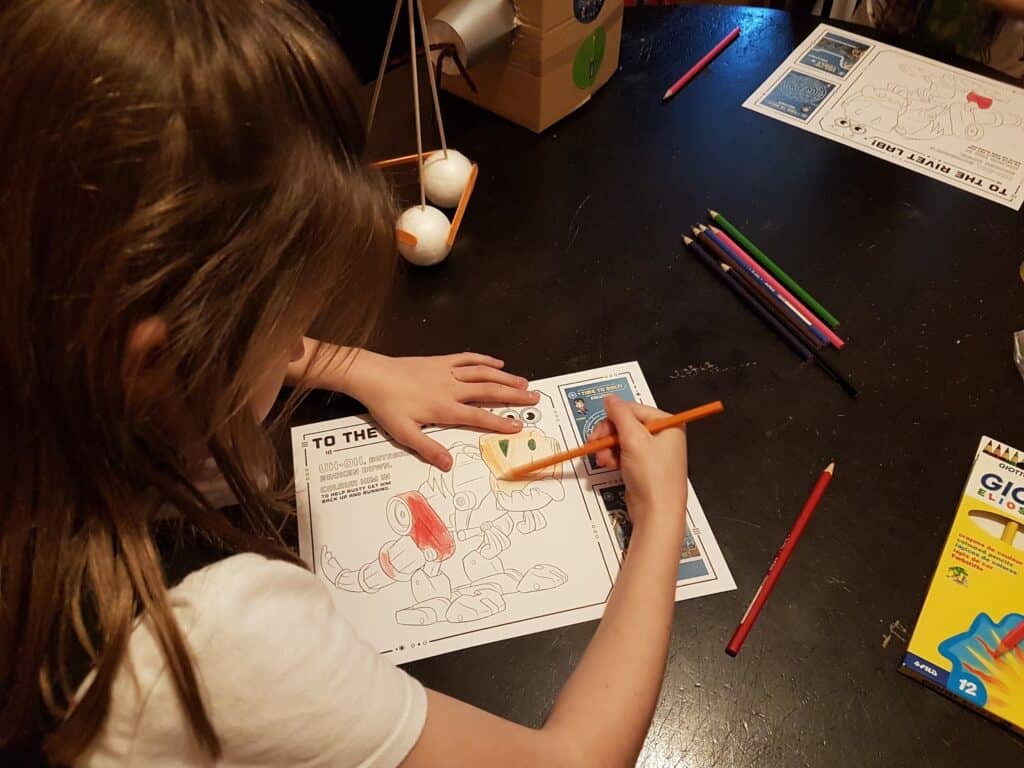 A child is colouring a robot on a printed worksheet at a dark table, using coloured pencils. Other coloured pencils, another worksheet, and a box of crayons are also on the table. The child’s focus is on the colouring activity.