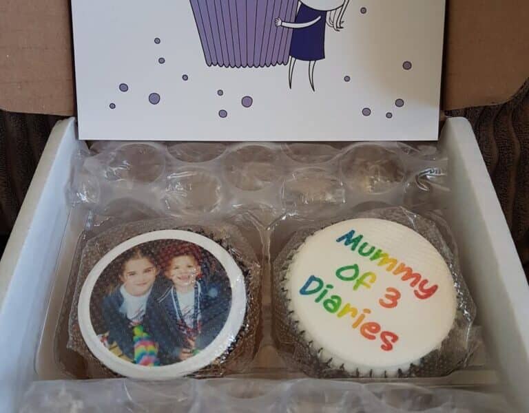 An open box contains two cupcakes—one with a photo of two children, the other with “Mummy Of 3 Diaries” in colourful icing—and a card featuring a cartoon girl holding a giant purple cupcake, all nestled in bubble wrap for protection.