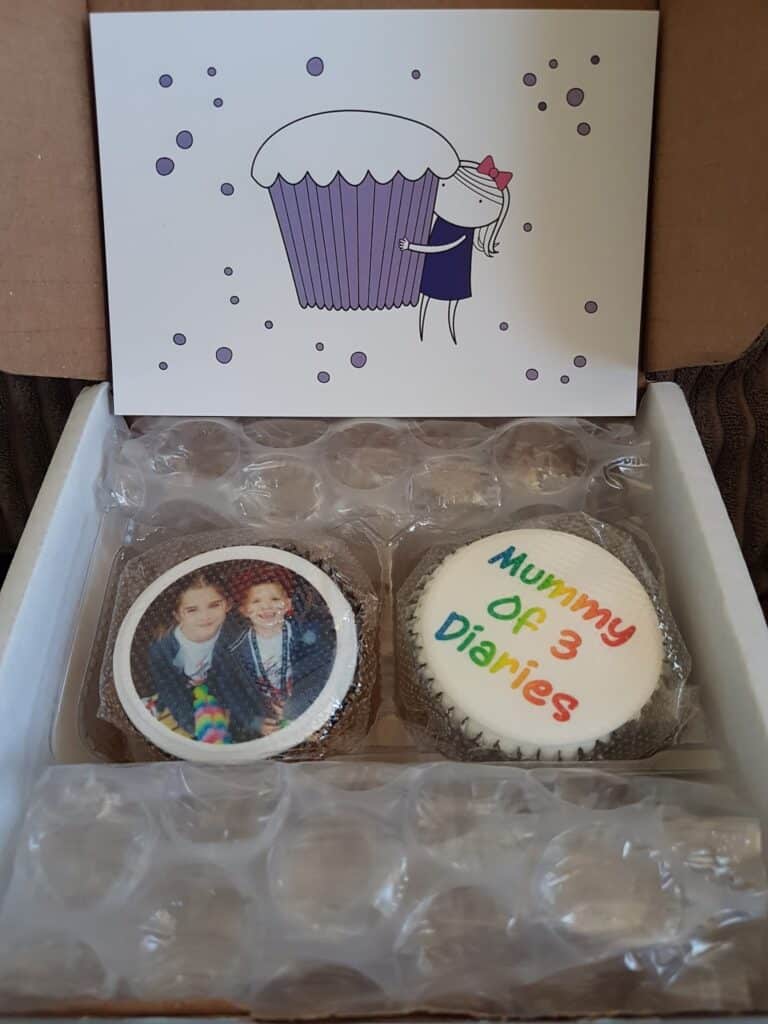 An open box with bubble wrap contains two cupcakes. One cupcake has a photo of two children printed on the icing. The other cupcake has “Mummy Of 3 Diaries” written in colourful letters. Behind them is a card showing a cartoon girl holding a giant cupcake.