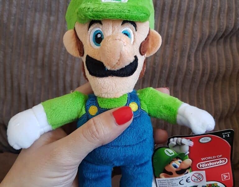 A hand with red painted nails holds a plush Luigi toy from Nintendo’s Mario series. Luigi wears his signature green hat with an “L”, green shirt, blue dungarees, and brown shoes. A tag with Luigi’s image and “World of Nintendo” branding is attached.