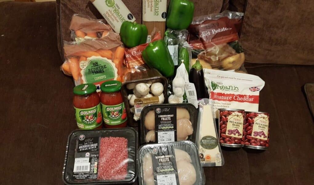 A variety of groceries are displayed on a settee, including green peppers, sweet potatoes, carrots, mushrooms, onions, pasta, two jars of tomato sauce, mature cheddar cheese, red kidney beans, chicken breast fillets, diced beef, and a pack of mixed apples.