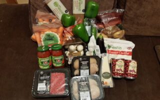 A variety of groceries are displayed on a settee, including green peppers, sweet potatoes, carrots, mushrooms, onions, pasta, two jars of tomato sauce, mature cheddar cheese, red kidney beans, chicken breast fillets, diced beef, and a pack of mixed apples.