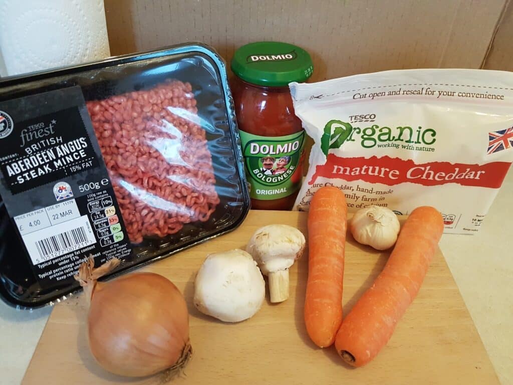 A wooden board holds a whole onion, two carrots, and two mushrooms. Behind them are a pack of raw beef mince, a jar of Dolmio Bolognese sauce, and a block of Tesco mature cheddar cheese. All items are arranged on a kitchen worktop.