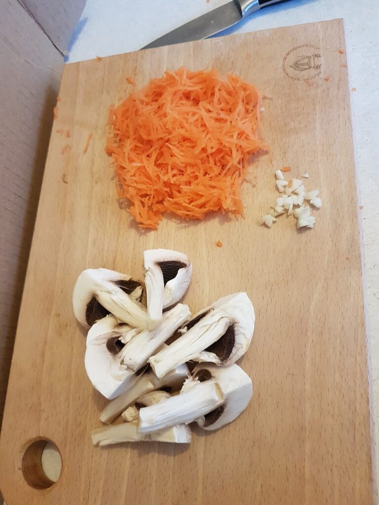 A wooden chopping board with three types of chopped vegetables: a pile of grated carrot, a small amount of finely minced garlic, and several quartered white mushrooms. A knife is partially visible at the top of the chopping board.