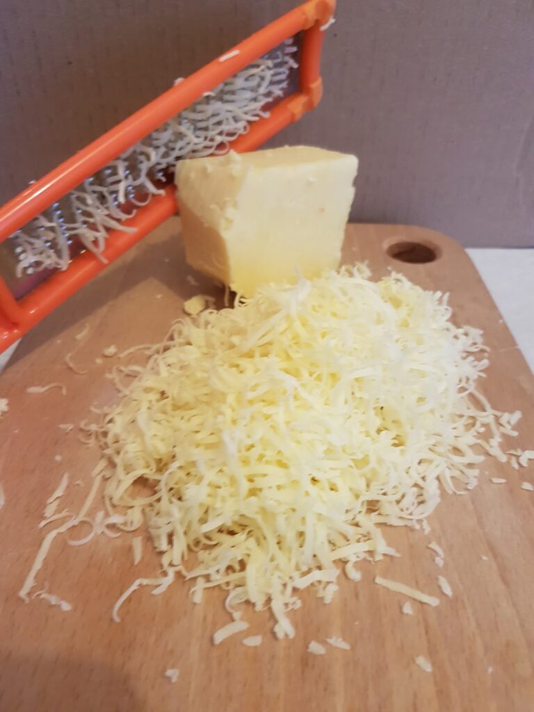 A wooden chopping board holds a pile of finely grated cheese, a wedge of ungrated cheese, and an orange-handled grater with some cheese remnants stuck to it. The background is plain and neutral.