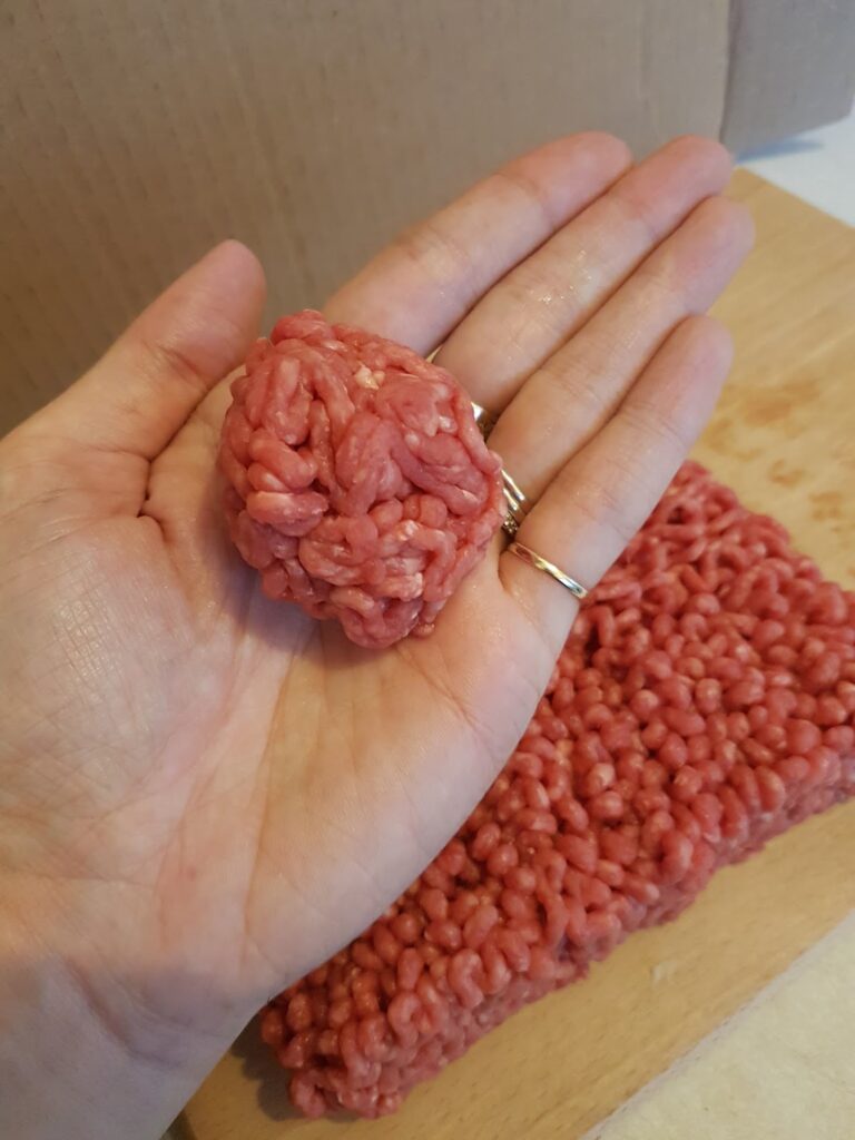 A hand holds a small, round ball of raw minced beef above a wooden surface. Below, a larger portion of raw minced beef rests on a chopping board. The person’s hand is open, displaying the meatball, with a gold ring on their finger.
