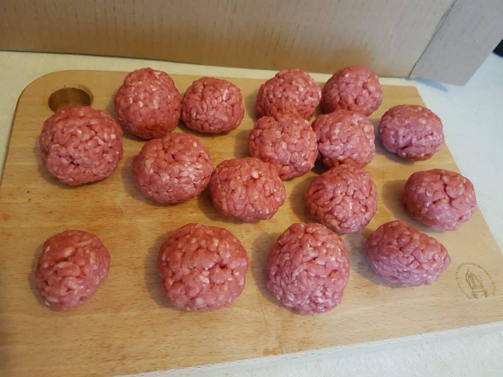 Several raw meatballs made from minced meat are neatly arranged on a light wooden chopping board. The meatballs vary slightly in size and shape. The background includes a cardboard box and a light-coloured surface.