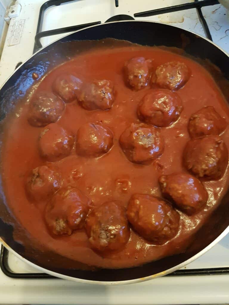 A frying pan on a hob contains several large meatballs simmering in a thick red tomato sauce. The meatballs are evenly coated with the sauce, and steam is slightly visible, suggesting they are being cooked.